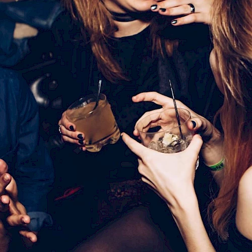 People are socializing, holding drinks, making gestures, and appearing to enjoy a lively atmosphere.