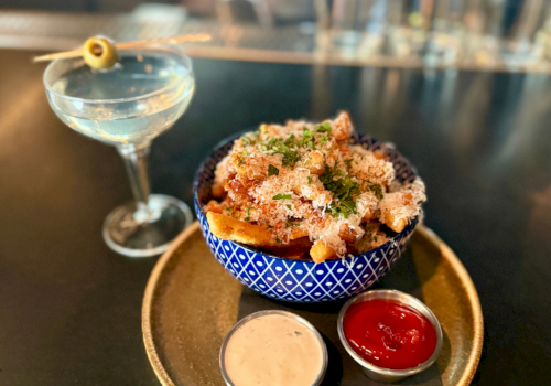 A blue bowl of bread topped with herbs and parmesan sits with two dipping sauces and a clear martini glass drink.