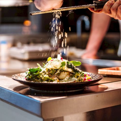 A chef is grating cheese over a beautifully plated dish in a professional kitchen setting, with focused attention on the food.