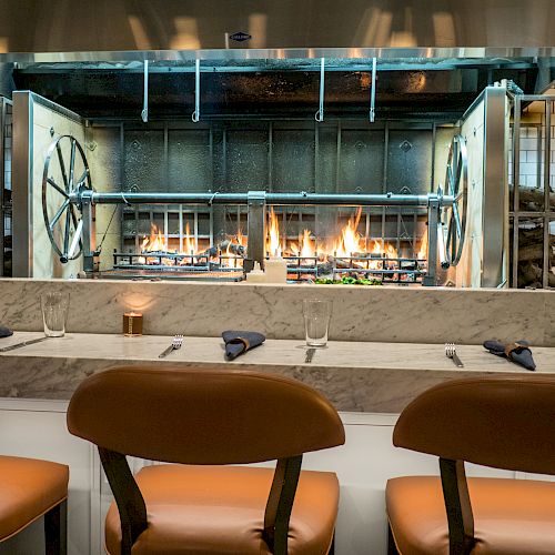 The image shows a restaurant kitchen with a grill and fire burning. In the foreground are bar stools and a counter set with utensils and glasses.