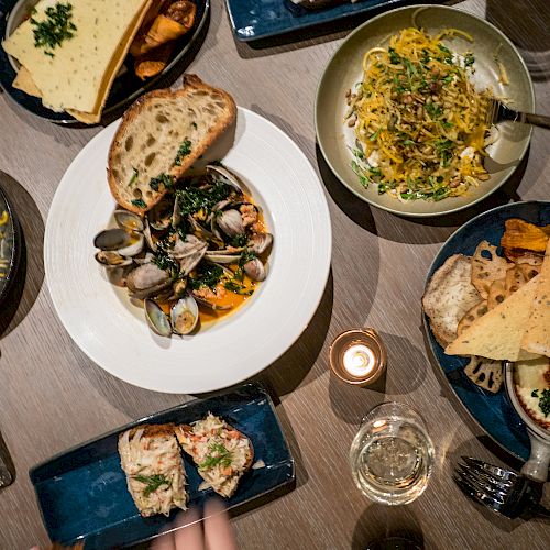 A table set with various dishes including pasta, bread, and seafood, surrounded by hands holding utensils and glasses.