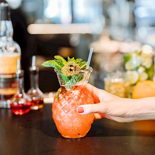 A person holding a tropical cocktail in a pineapple-shaped glass, garnished with mint and a flower. Various liquor bottles are in the background.