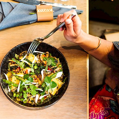 A person is holding a fork over a bowl of salad with various greens, seeds, and vegetables on a wooden table adorned with napkins and a napkin ring.