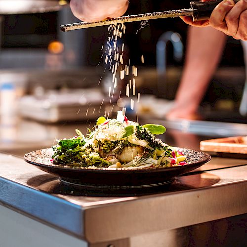 A chef grates cheese over a plated dish in a kitchen.