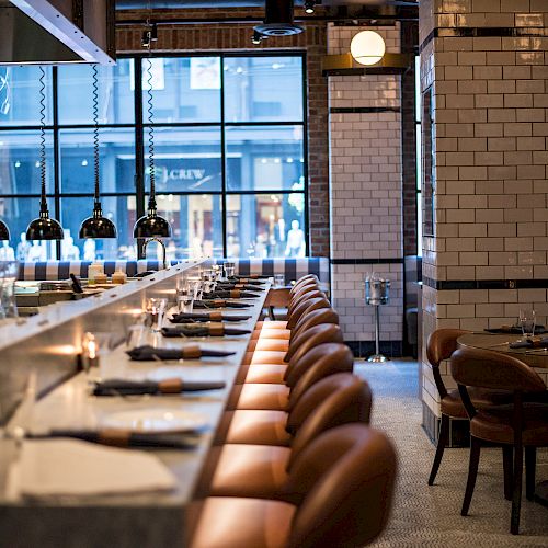 A modern, stylish restaurant interior featuring a long marble bar with brown leather stools, white tiled walls, and pendant lighting, ending the sentence.