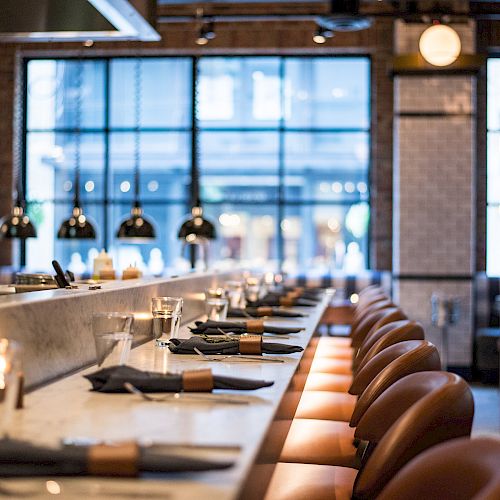 This image shows the interior of a modern, well-lit restaurant with a long counter, bar stools, and place settings ready for guests.