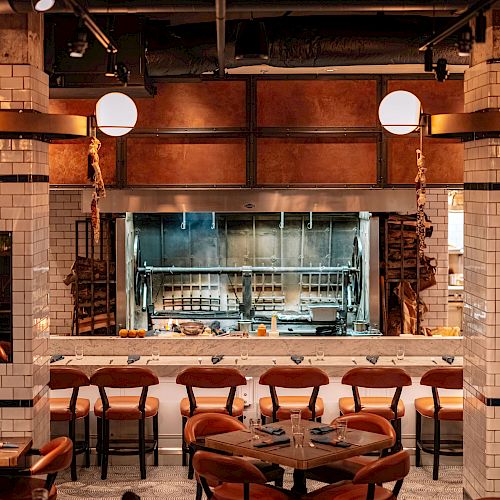 The image shows a cozy restaurant interior with wooden tables, brown chairs, white tiled walls, hanging lights, and an open kitchen at the back.