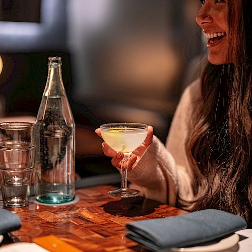 A woman is sitting at a table in a restaurant, holding a drink and smiling, with a bottle of water, a glass, and a menu on the table.