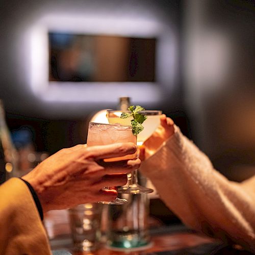 Two people clinking glasses in a warmly lit setting, possibly a restaurant or bar, with menus and napkins on the table.