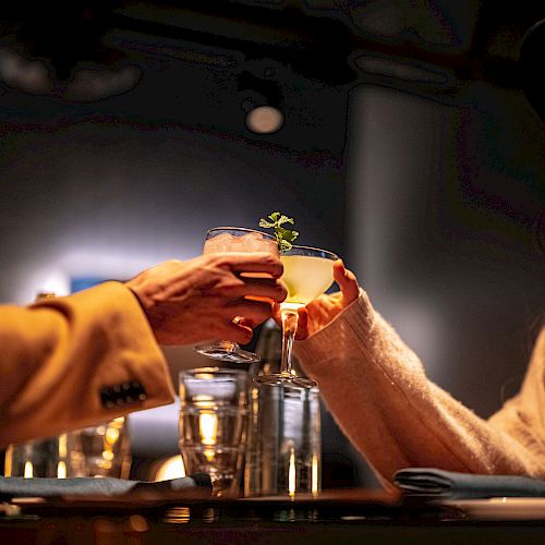 Two people are seated at a table, clinking their drinks in a toast, in a dimly lit setting. There are water glasses on the table.