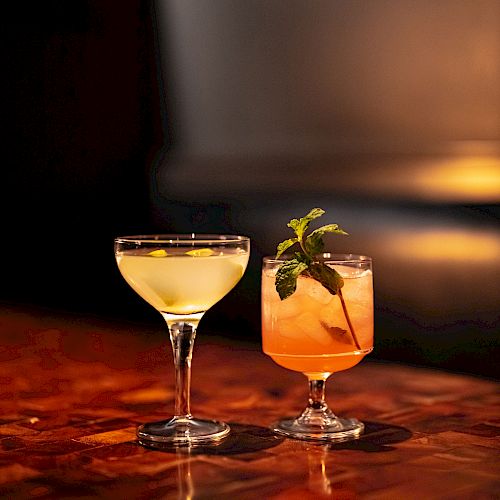 Two cocktails on a polished wooden table. One is light yellow in a coupe glass, and the other is peach-colored with mint garnish in a rocks glass.