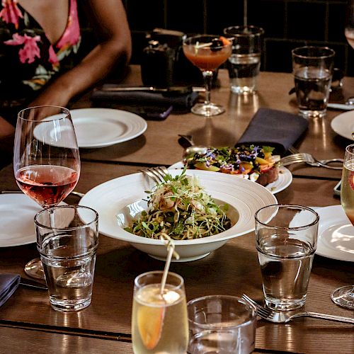 A table with various drinks, a bowl of salad, plates, glasses, and a person in a floral dress seated at the table.