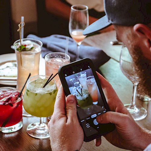 A person is taking a photo of various colorful drinks on a table. Others are seated around the table, partially visible in the background.