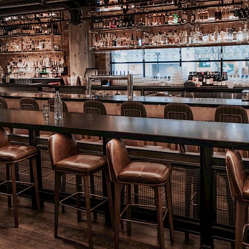 The image shows an empty bar with high stools, a long counter, and shelves stocked with various bottles of alcohol behind it.