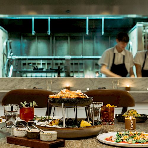 A restaurant setting with various dishes and drinks on a table in the foreground, while two chefs work in an open kitchen in the background.