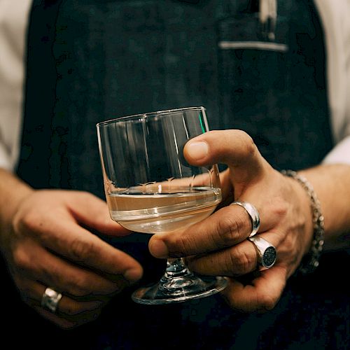 A person wearing a dark apron is holding a glass of light-colored liquid, with several rings on their fingers and a pen in the apron pocket.