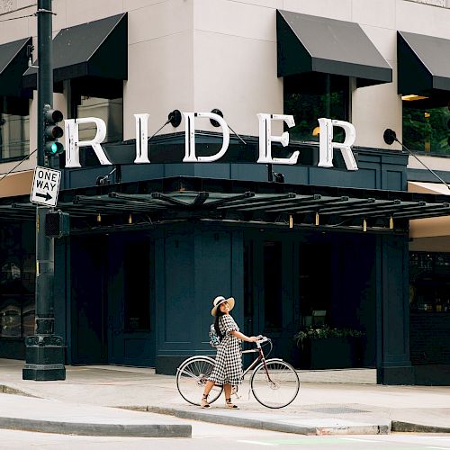 A person walks their bike on a city street in front of a building with the large sign "RIDER" and black awnings above the windows.