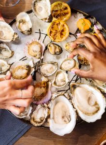 Three people are sharing a platter of oysters with grilled lemons, served on a bed of ice, accompanied by various dipping sauces.