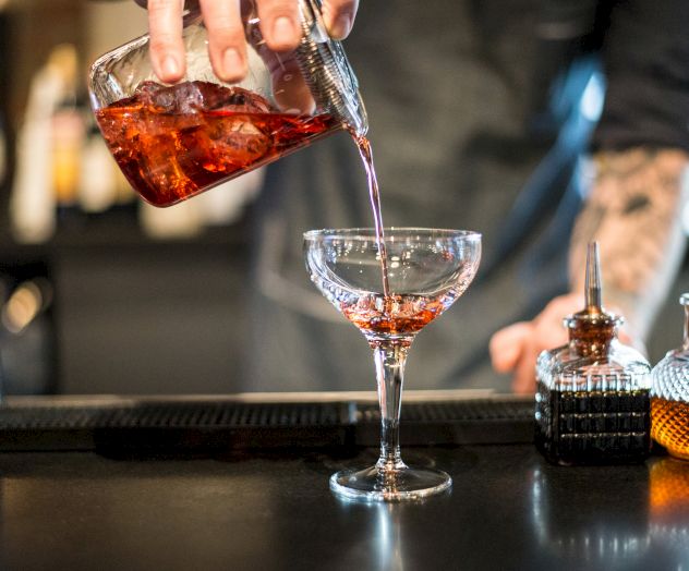 A bartender is pouring a red cocktail from a mixing glass into a stemmed glass on a bar, next to various bottles and tools.