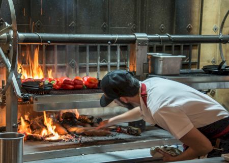 A chef, working in an industrial kitchen, tends to a fiery grill with vegetables on it, ensuring proper cooking.