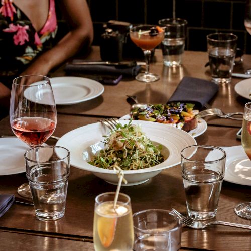 A group is gathered around a table with plated food, drinks, and glasses, suggesting a dining setting. A bowl of salad is in the center of the table.