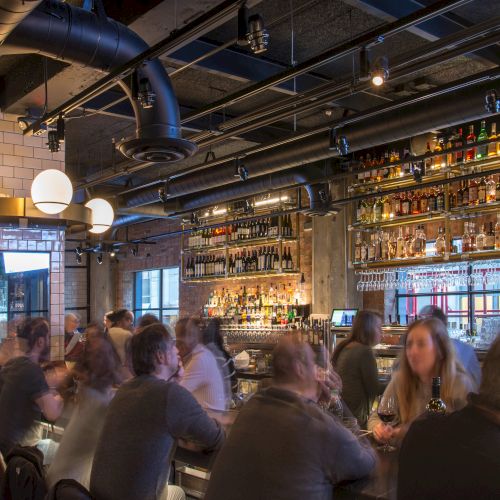 People are sitting and socializing at a busy bar with a well-stocked shelf of liquor bottles in the background.