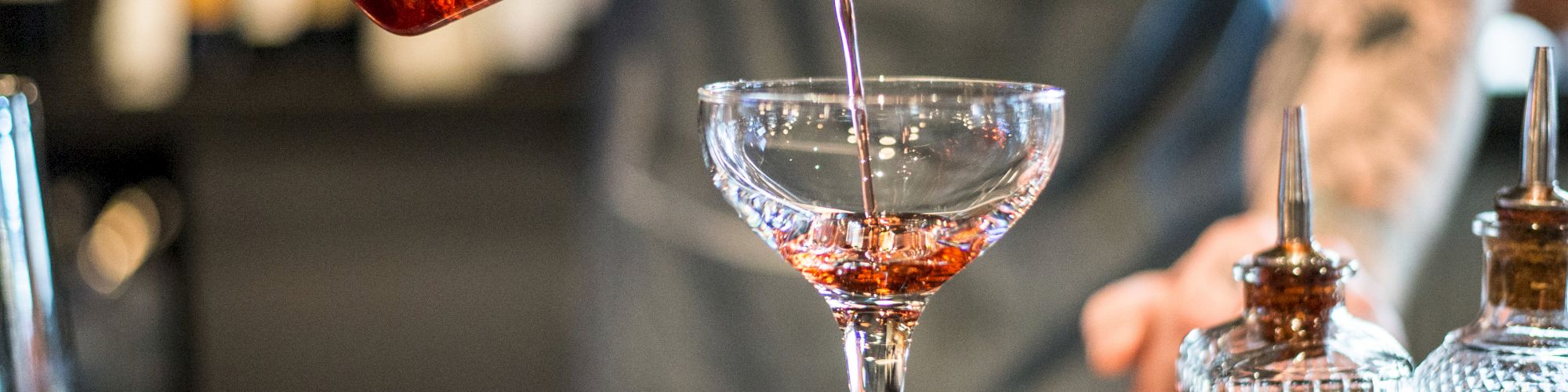 A bartender pours a red cocktail from a mixing glass into a coupe glass, with bitters bottles placed on the counter.