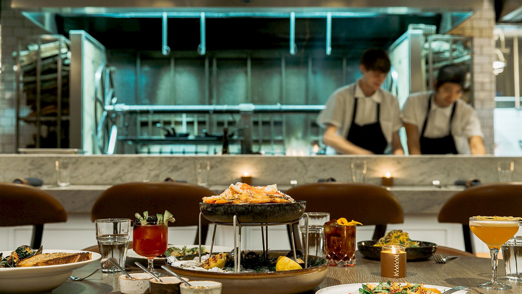 A restaurant scene shows a table with various dishes and drinks, with two chefs working in an open kitchen in the background.