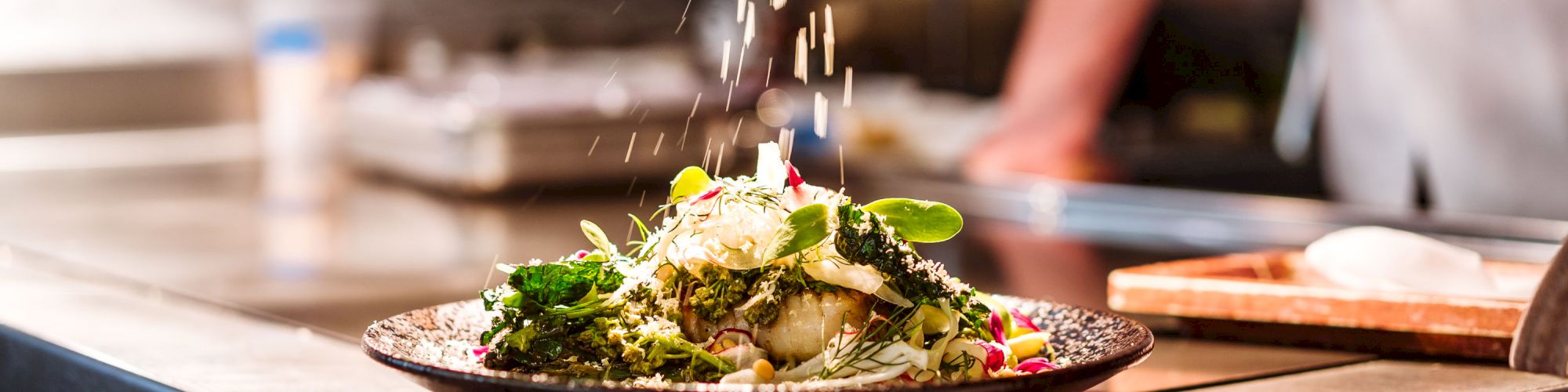 A person is grating cheese over a plated dish with greens and vegetables in a kitchen setting.