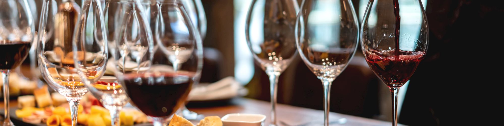 A person is pouring red wine into glasses, surrounded by a table set with wine glasses and plates of assorted cheeses and charcuterie.