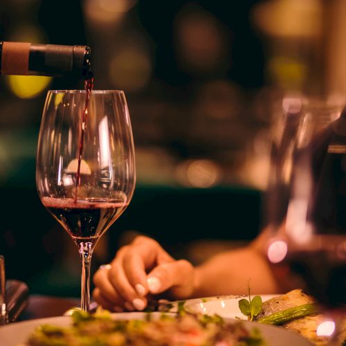 A person pours red wine into a glass on a dinner table, with another glass and a plate of food visible.