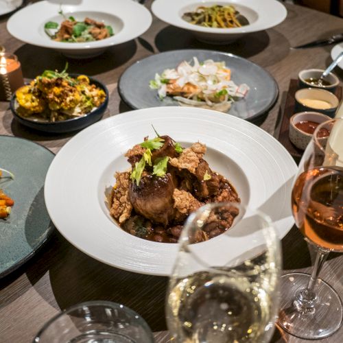 A table filled with various gourmet dishes and wine glasses, featuring a central dish of meat and greens, surrounded by plates of pasta, salads, and sides.