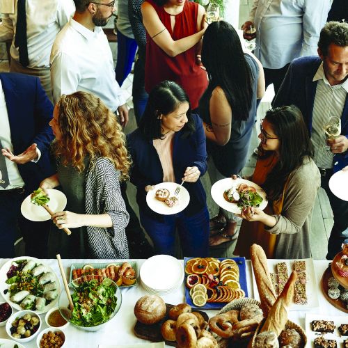 A group of people at a social gathering, holding plates and conversing, with a table full of various foods and drinks in the foreground.