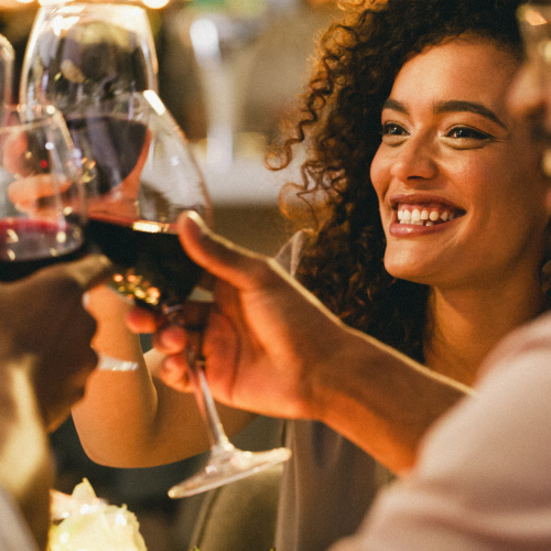 A group of people clinks wine glasses together in a celebratory toast, with a woman smiling warmly at the center of the scene.