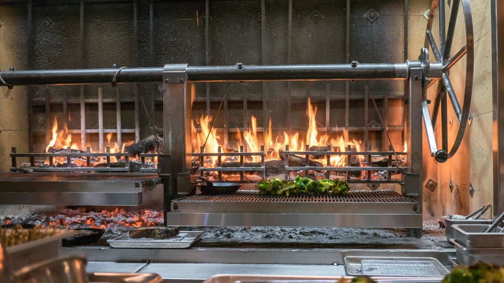 A grill with flames and vegetables cooking over an open flame, set in a professional kitchen environment.