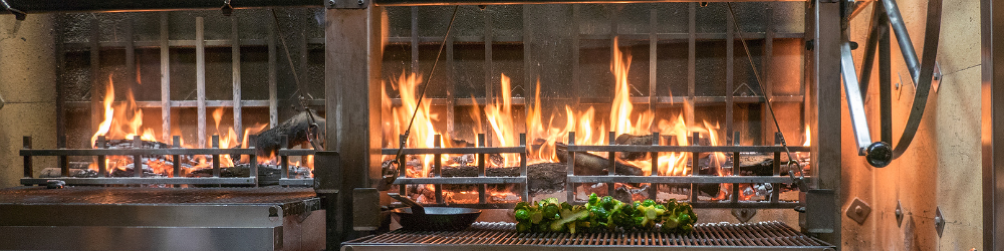 A grill with flames and vegetables cooking over an open flame, set in a professional kitchen environment.