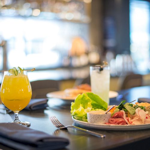 The image shows a dining table with plates of appetizers, salads, and two colorful drinks in a restaurant setting.