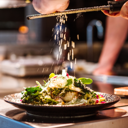 A chef grates cheese over a plated dish with greens, herbs, and garnishes in a kitchen setting.