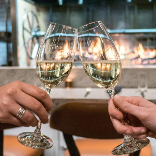 Two people clink wine glasses in front of a cozy fireplace, creating a warm and celebratory atmosphere.