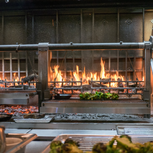 An open flame grill with cooking surfaces and vegetables, featuring a wheel mechanism, in a professional kitchen setting.