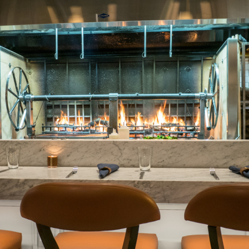 An open kitchen with a large grill, flames visible, and a marble countertop, set with chairs, napkins, and glasses in the foreground.