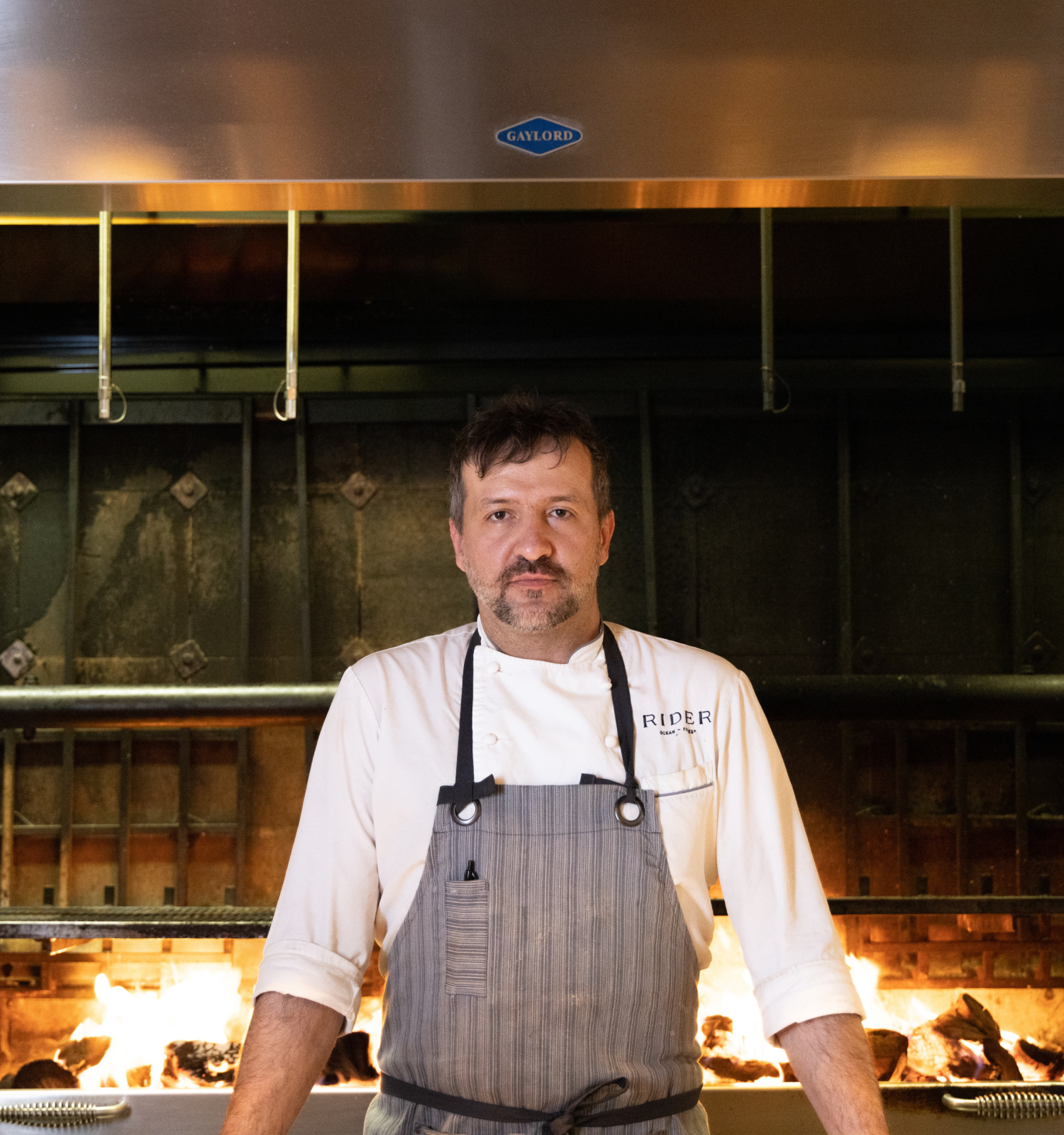 A chef stands in front of an open flame grill in a kitchen, wearing a white chef jacket and an apron, with metal wheels on either side.