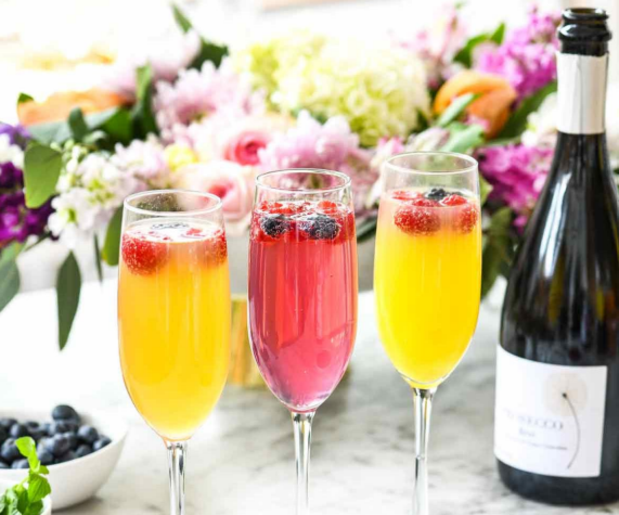 Three glasses of mimosas with fruit, a bottle of Prosecco, a bowl of blueberries, and a colorful bouquet of flowers on a marble surface.