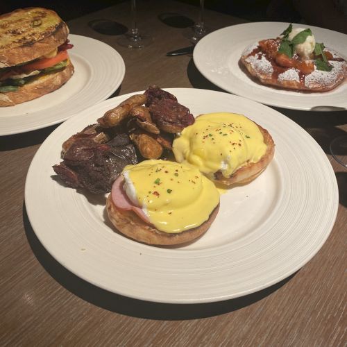 The image shows a meal with dishes including Eggs Benedict, a sandwich, and a plate with toppings, accompanied by drinks in the background.