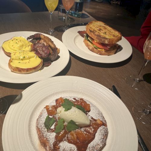 Three dishes are served on a table with glasses of beverages. They include a dessert, possibly pancakes or waffles, and savory breakfast items.