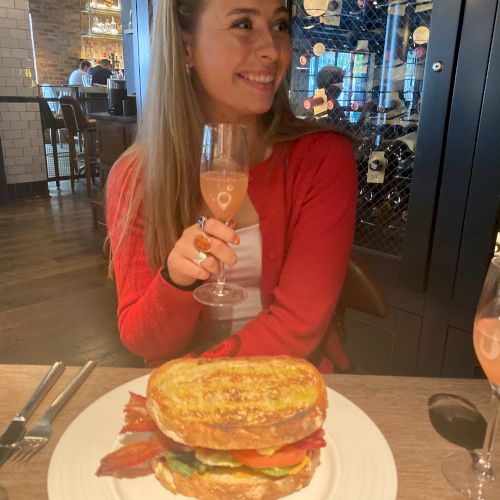 A person sitting at a restaurant holding a drink, with a large sandwich on a plate in front of them, smiling and looking to the side.