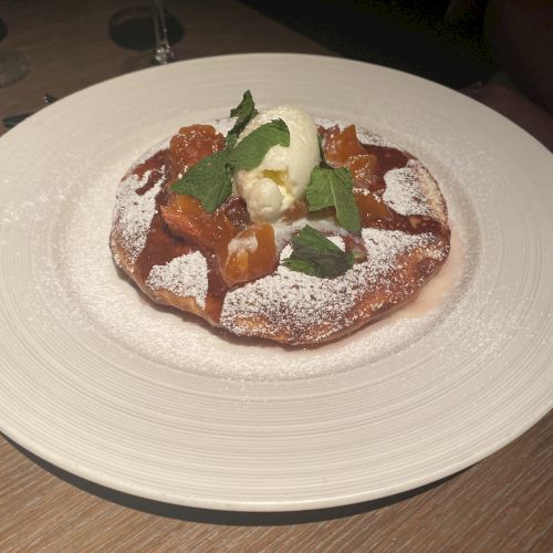 This image shows a dessert topped with powdered sugar, fruit pieces, a scoop of ice cream, and a mint leaf, served on a white plate.