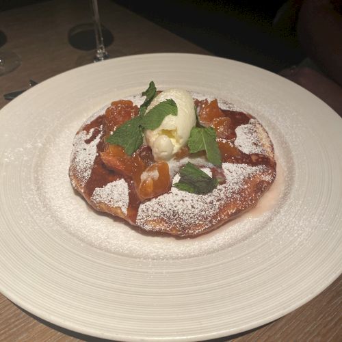 The image shows a dessert with fruit, ice cream, and mint leaves on top of powdered sugar, served on a white plate.