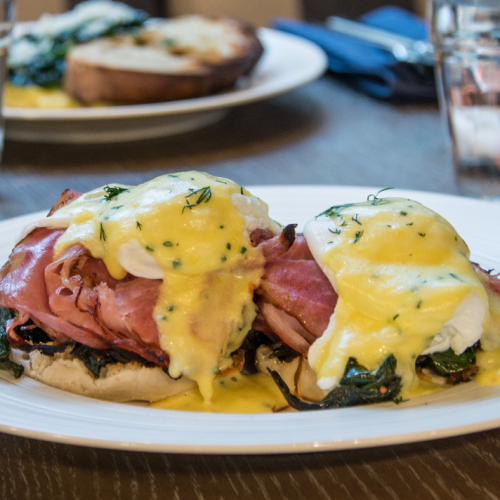 The image shows a plate of Eggs Benedict topped with hollandaise sauce, ham, and sautéed spinach on an English muffin. Another plate is in the background.