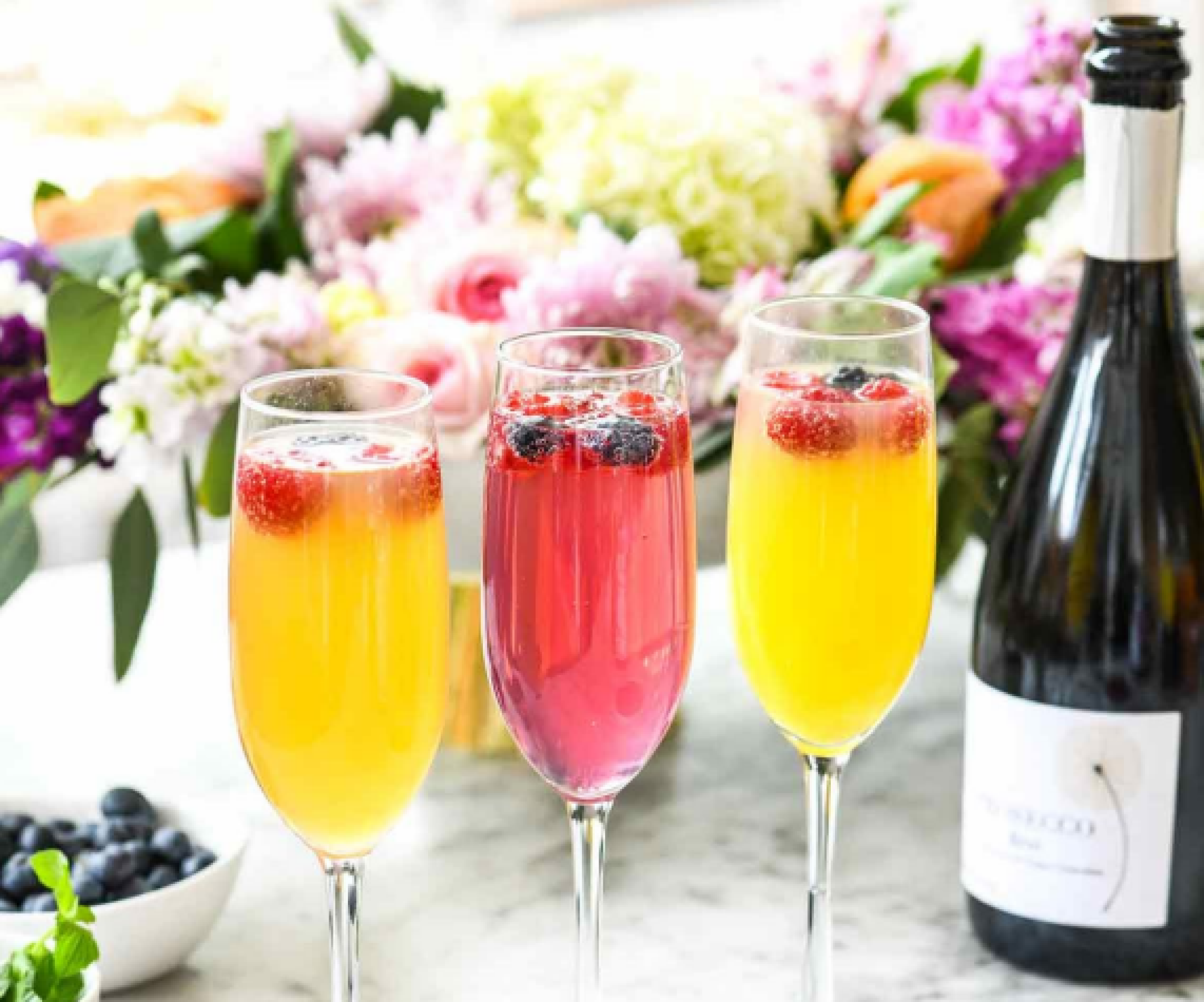 Three champagne glasses with colorful drinks and berries, a bottle of prosecco, a bowl of blueberries, and a flower arrangement in the background.